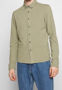 Olivově zelená košile s dlouhým rukávem a límcem s knoflíky, přední knoflíky a malým logem; přiřazena s modrými denimovými džíny.