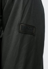 Jachetă neagră din nailon, cu o țesătură texturată, având o etichetă dreptunghiulară proeminentă din piele neagră inscripționată cu "Barbour International".