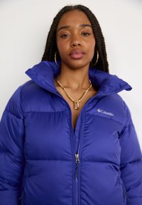 Jachetă puffer Columbia în violet vibrant, cu un guler înalt, închidere cu fermoar și buzunare laterale cu un model matlasat.