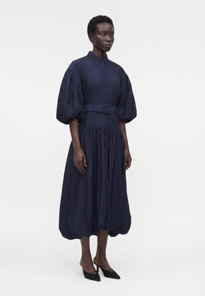 Γυναίκα φοράει ένα μέτριο φόρεμα σε ναυτικό μπλε με ζώνη, φουσκωτά μανίκια και ραμμένη φάσα, συνδυασμένο με μαύρες γόβες slingback με μυτερή μύτη.