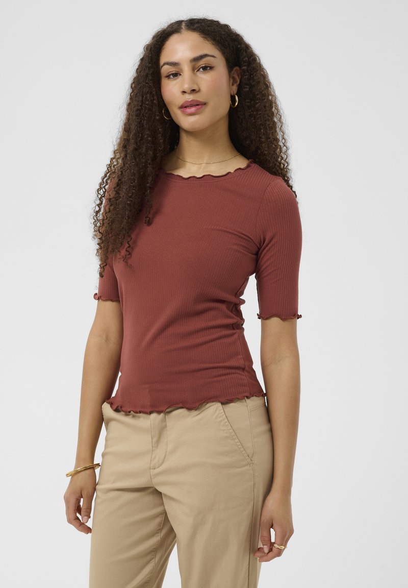 Femme aux longs cheveux bouclés portant un haut côtelé rouge rouille avec des bords festonnés et un pantalon beige, debout devant un fond blanc uni.
