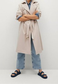 Beige trenchcoat met een ceintuur in de taille en een dubbelgeknoopte ontwerp, gecombineerd met lichtblauwe denim wijde pijpen jeans en marineblauwe instapsandalen.