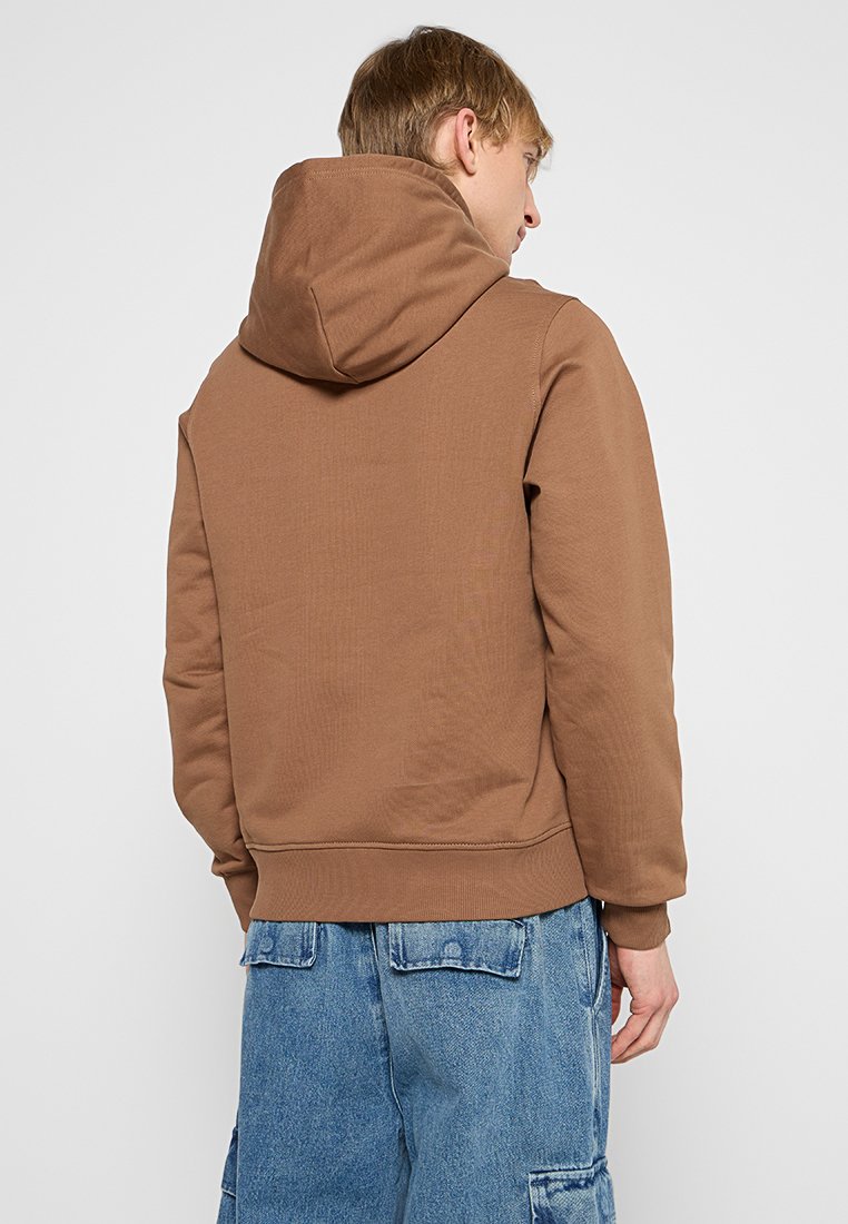 Brązowa bluza z kapturem, z prążkowanym wykończeniem na dole i mankietach, charakteryzująca się przestronnym kapturem oraz gładkim, teksturowanym materiałem; noszona z luźnymi dżinsowymi szortami.
