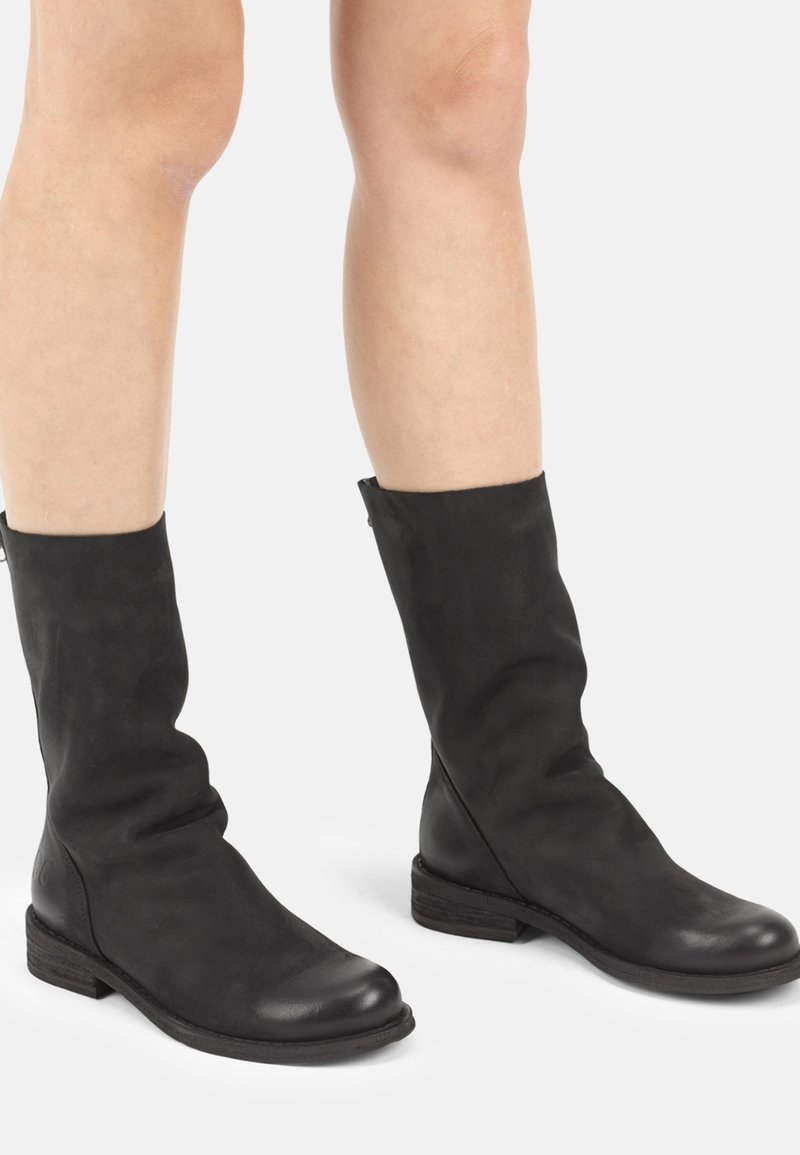 Par de botas negras de cuero hasta la mitad de la pantorrilla, usadas sobre piernas desnudas, con tacones bajos apilados y punteras redondeadas sobre un fondo claro y liso.