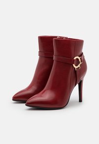 Stivaletti alla caviglia in pelle rossa con punta affilata, tacco sottile e una fascia con un accento in metallo dorato rotondo. Tessuto liscio e design elegante.