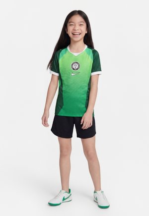 Niña joven sonriendo, lleva una camiseta de fútbol con degradado verde y logo, pantalones cortos negros y zapatillas blancas con detalles verdes, de pie sobre un fondo blanco.