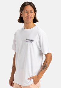 Camiseta blanca de manga corta hecha de algodón, que presenta un parche del logo "Quiksilver" en azul y negro en el área del pecho izquierdo.