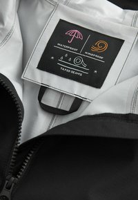 Interior de una chaqueta negra impermeable con un forro blanco, que presenta una etiqueta con símbolos de impermeabilidad y resistencia al viento, así como costuras selladas.