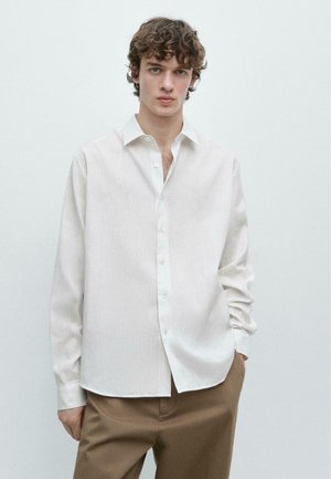 Junger Mann mit lockigem Haar, der ein locker sitzendes weißes Hemd mit Knöpfen und braune Hose trägt, vor einem schlichten hellen Hintergrund stehend.