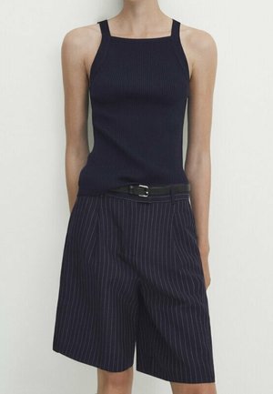 Nainen, joka pitää yllään tummansinistä ribbineuletakkia ja tummansinisiä, mustilla vyöllä varustettuja polvipituisia raidallisia shortseja, seisoo vaaleaa yksinkertaista taustaa vasten.