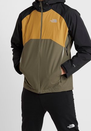 Man draagt een North Face jas in drie kleuren met zwarte mouwen, mosterdgele borst en olijfgroene romp, handen in de zakken, zwarte broek.