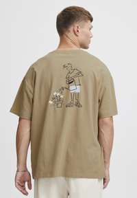 Khaki bomulds t-shirt med korte ærmer og oversize pasform. Har en sort linjetegning af en person, der vander planter, på ryggen.