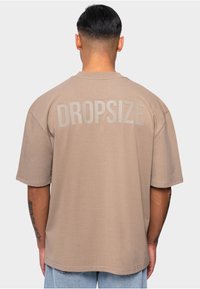 T-shirt oversize marron à manches courtes, arborant le mot "DROPSIZE" en grandes lettres gris clair au dos. Texture lisse.