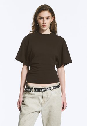 Tânără cu păr ondulat, purtând un tricou negru mulat cu mâneci scurte și pantaloni bej deschis, cu o curea neagră cu ținte, stând în picioare.