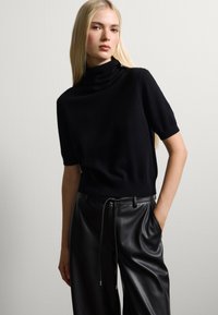 Maglione nero a maniche corte con collo alto realizzato in materiale morbido, abbinato a pantaloni in pelle neri a vita alta con una cintura regolabile.