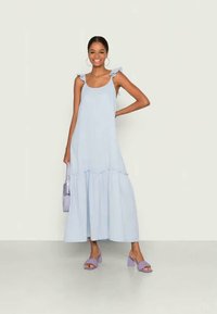 Šviesiai mėlynas maksi suknelė su voliotais petnešomis, sluoksniuota išvaizda ir laisvai krentanti siluetu. Sujungta su levandų spalvos bloko kulno sandalais ir mažyte rankine.