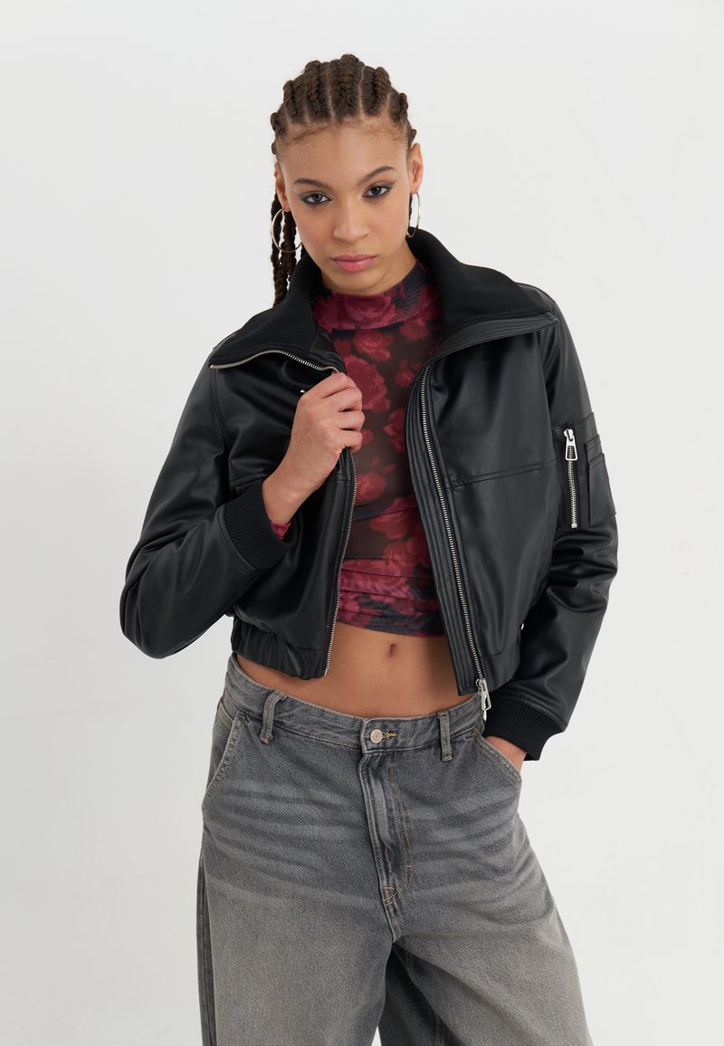 Black faux leather cropped jacket with a zippered front, ribbed collar, and side pockets, paired with a patterned top and gray high-waisted jeans.