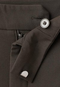 Gros plan sur la ceinture d’un pantalon en tissu foncé avec un bouton argenté, une fermeture éclair partiellement visible et un petit fermoir argenté sur le rabat.