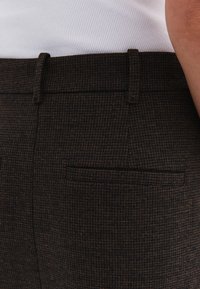 Pantalones marrones con un patrón, con un tejido texturizado, que presentan un discreto bolsillo trasero y un corte ajustado, mostrados desde un ángulo lateral.