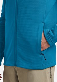 Chemise à manches longues zippée bleue avec un motif texturé, comprenant des poches latérales et un cordon de serrage au bas. Tissu doux et léger.