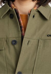 Olivově zelená bunda s límcem, černými knoflíky a dvěma kapsami. Přes ni je navrstvená kostkovaná flanelová košile a hořčicově oranžový svetr.