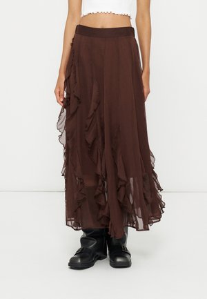 Jupe longue marron transparente avec des détails à volants portée avec des bottes noires à boucle et un top court blanc.