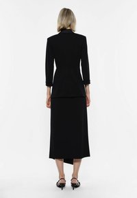 Blazer nero sartoriale con maniche a tre quarti, abbinato a una gonna lunga coordinata. Tessuto morbido, design elegante, minimalismo nei dettagli e scarpe con tacco alto.