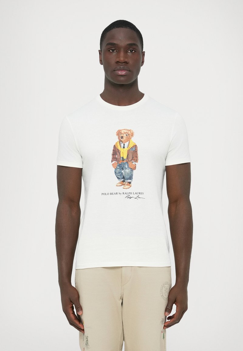 T-shirt branca de algodão com uma ilustração colorida de um urso com um casaco castanho e lenço amarelo. Texto abaixo diz "POLO BEAR por RALPH LAUREN."