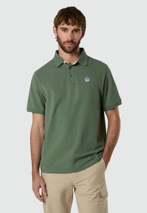 Mann trägt grünes kurzärmliges Polohemd mit blau-weißem Logo, steht mit einer Hand in der beige Hose Tasche vor einfachem Hintergrund.
