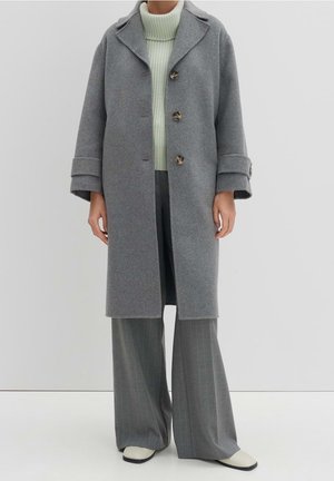 Persoon die een grijze wollen jas draagt over een lichtgroene coltrui, brede grijze broek met fijne strepen en witte schoenen tegen een effen achtergrond.