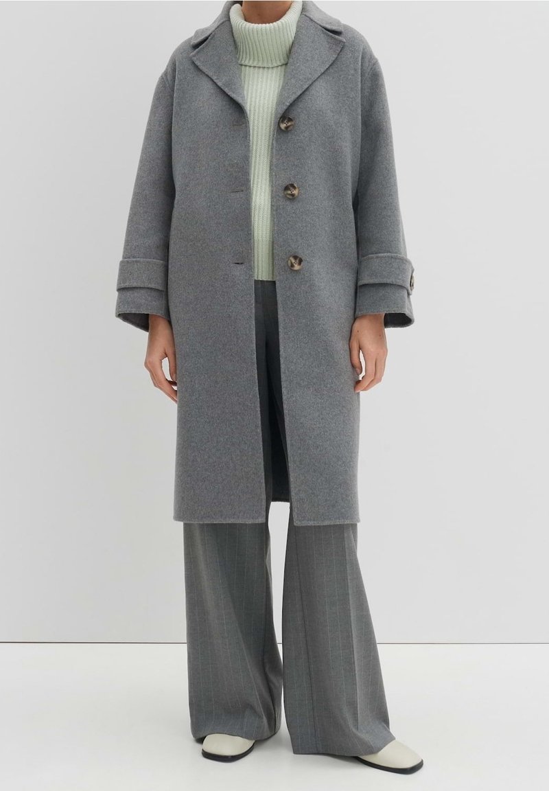 Personne portant un manteau en laine gris sur un pull à col roulé vert clair, un pantalon large gris à fines rayures et des chaussures blanches sur un fond uni.