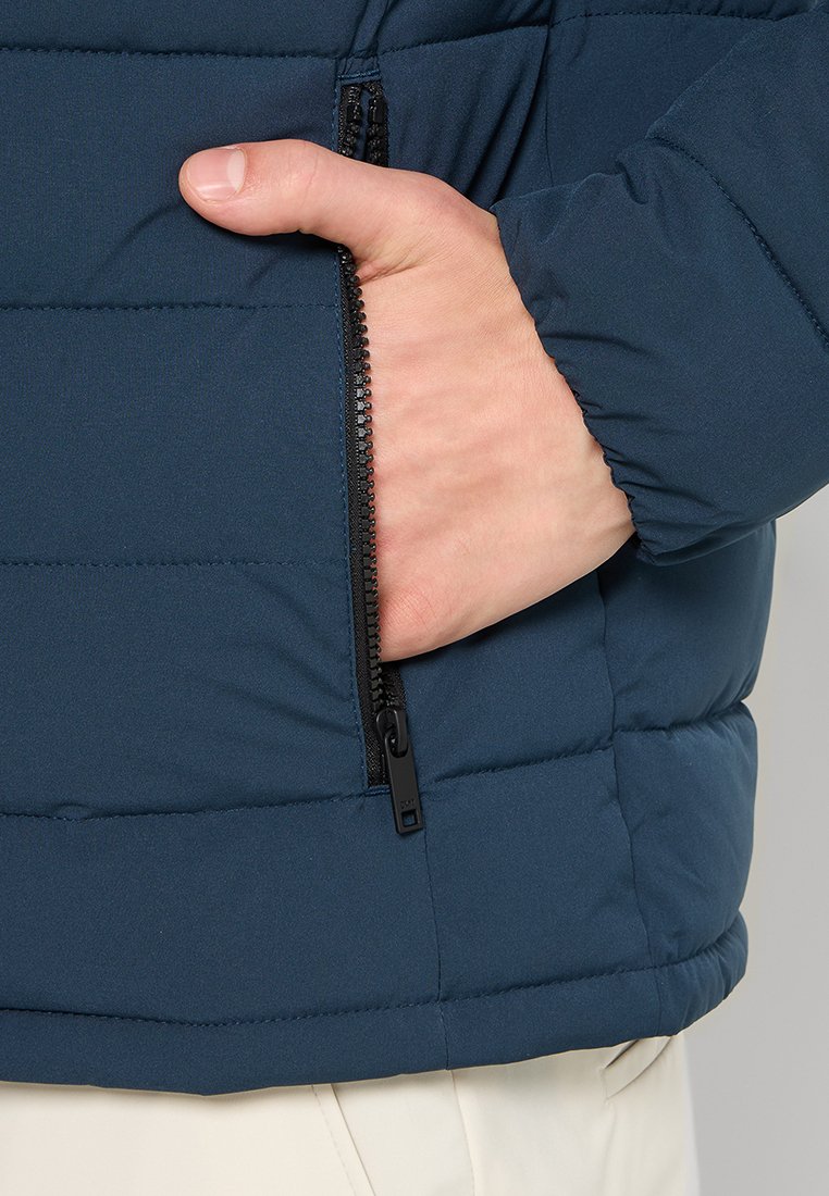 Padded jas in donkerblauw, met een ritszak aan de zijkant. Materiaal heeft een gladde textuur met gestikte panelen voor isolatie.