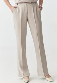 Pantaloni beige classici con gamba dritta, pieghe frontali pulite e tasche laterali, realizzati in un tessuto morbido. Scarpe non visibili.