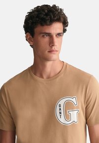 Camiseta de algodón color vainilla con un gran gráfico en blanco y negro que incluye la palabra "GANT" y una letra "G" prominente en el pecho.