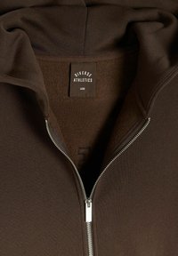 Brązowa bluza z kapturem na zamek z miękkim wnętrzem. Posiada zamek błyskawiczny, dużą metkę rozmiarową i prosty design bez ozdób.