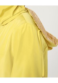 Chaqueta impermeable amarilla con un forro interior texturizado, que cuenta con capucha, puños elásticos y un cierre de cremallera en el cuello.