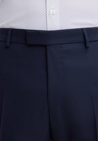 Pantaloni sartoriali blu marino con design a davanti piatto. Presentano passanti per cintura e tessuto finemente strutturato. Indossati con una camicia chiara e a fantasia.