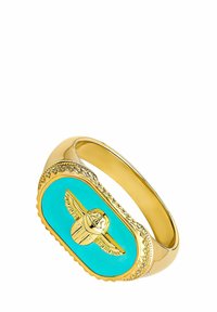 Pulsera en tono dorado que presenta una piedra ovalada de turquesa con un emblema alado incrustado. Bordes texturizados con detalles decorativos alrededor de la piedra.