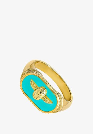 Pulsera en tono dorado que presenta una piedra ovalada de turquesa con un emblema alado incrustado. Bordes texturizados con detalles decorativos alrededor de la piedra.