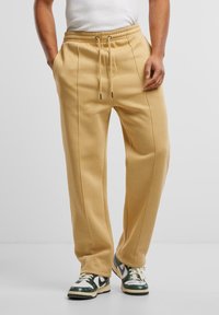 Beige katoenen sweatpants met een elastische tailleband, zijzakken en naaddetails; gedragen met groen-witte sneakers op een neutrale achtergrond.