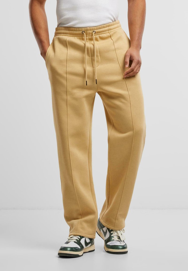 Beige katoenen sweatpants met een elastische tailleband, zijzakken en naaddetails; gedragen met groen-witte sneakers op een neutrale achtergrond.