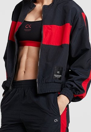 Atleet gekleed in een zwart-rood Calvin Klein trainingspak met een cropped jack, bijpassende sportbeha en hooggetailleerde broek met handen in de zakken.