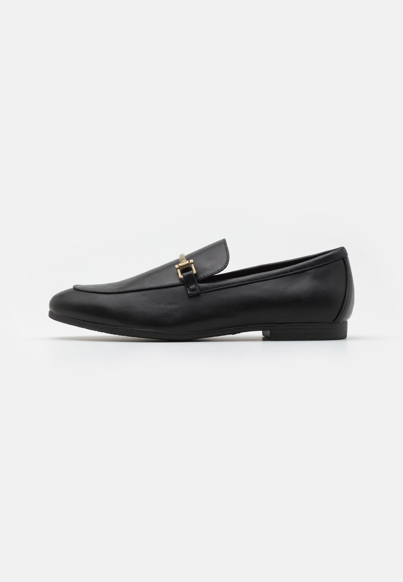 Mocassin noir en cuir à enfiler avec petit talon et détail de boucle métallique dorée sur fond blanc, vue de côté.