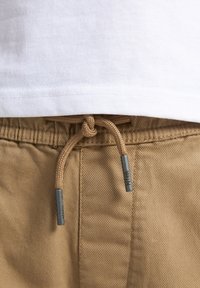 Pantaloni in cotone beige con superficie текстurizzata, dotati di un cordoncino annodato e aghetti in metallo argentato. Orlo della maglietta bianca visibile sopra.