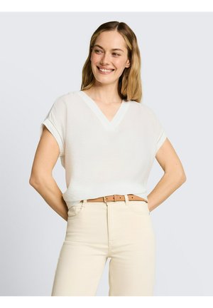 Uśmiechnięta kobieta o blond włosach, ubrana w biały top z krótkim rękawem i dekoltem w serek, włożony w beżowe spodnie z wysokim stanem oraz jasnobrązowy pasek.
