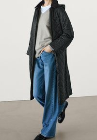 Zwarte doorgestikte lange jas met kraag, gedragen over een grijze trui en blauwe wijde jeans. Voorzien van drukknoopsluitingen en een gestructureerde afwerking.