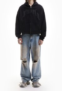 Personne portant un sweat à capuche noir zippé avec des lettres, un jean déchiré bleu clair et des baskets grises, se tenant devant un fond blanc.