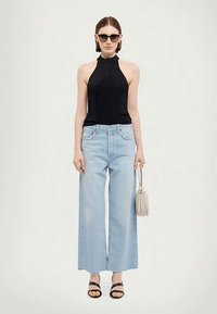 Kvinne iført svart halterneck-top, lyseblå vide jeans, svarte sandaler, mørke solbriller, holder en beige veske mot en ensfarget bakgrunn.