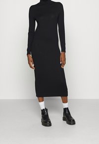 Schwarzes, figurbetontes Strickkleid mit hohem Kragen und langen Ärmeln. Kombiniert mit weißen Knöchelsocken und schwarzen Knöchelstiefeln. Glatte Textur.
