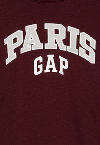 Felpa color marsala con lettere bianche in stile blocco che formano la scritta "PARIS GAP." Il testo è presentato in un arrangiamento curvo. Tessuto morbido.